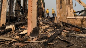 Two firefighters survey the remnants of a building destroyed by fire, surrounded by charred debris and ash.