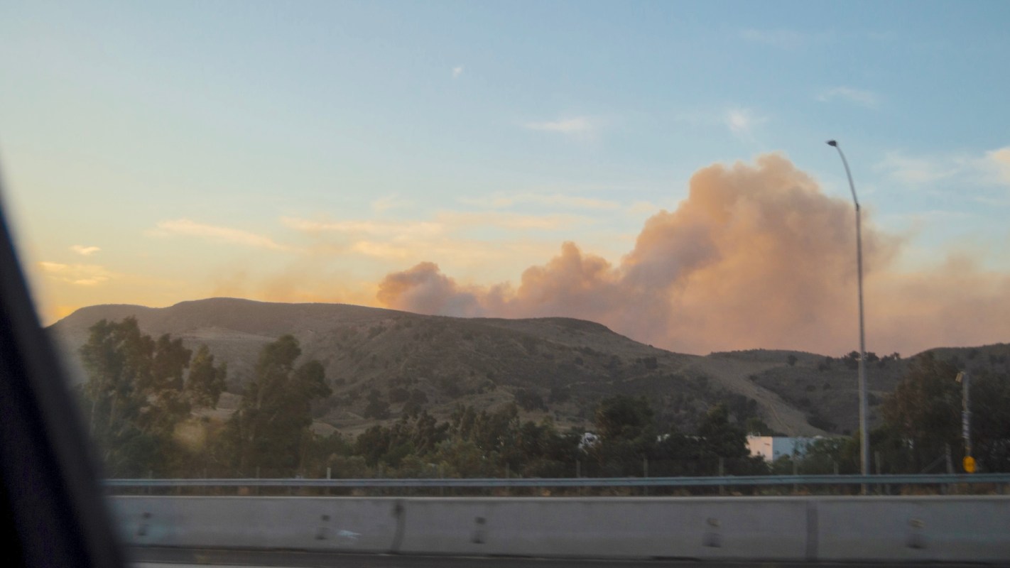 A landscape with hills in the background and smoke rising against a colorful sky at sunset.