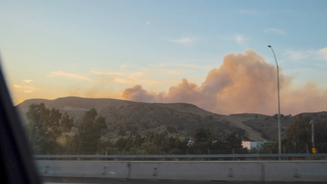 A landscape with hills in the background and smoke rising against a colorful sky at sunset.