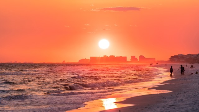 A vibrant sunset casts orange hues over a beach with silhouetted figures walking along the shoreline.