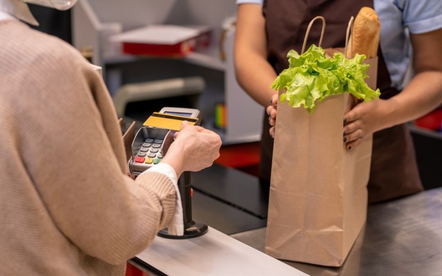 A customer is paying at a checkout counter while a cashier holds a paper bag with lettuce and a baguette.
