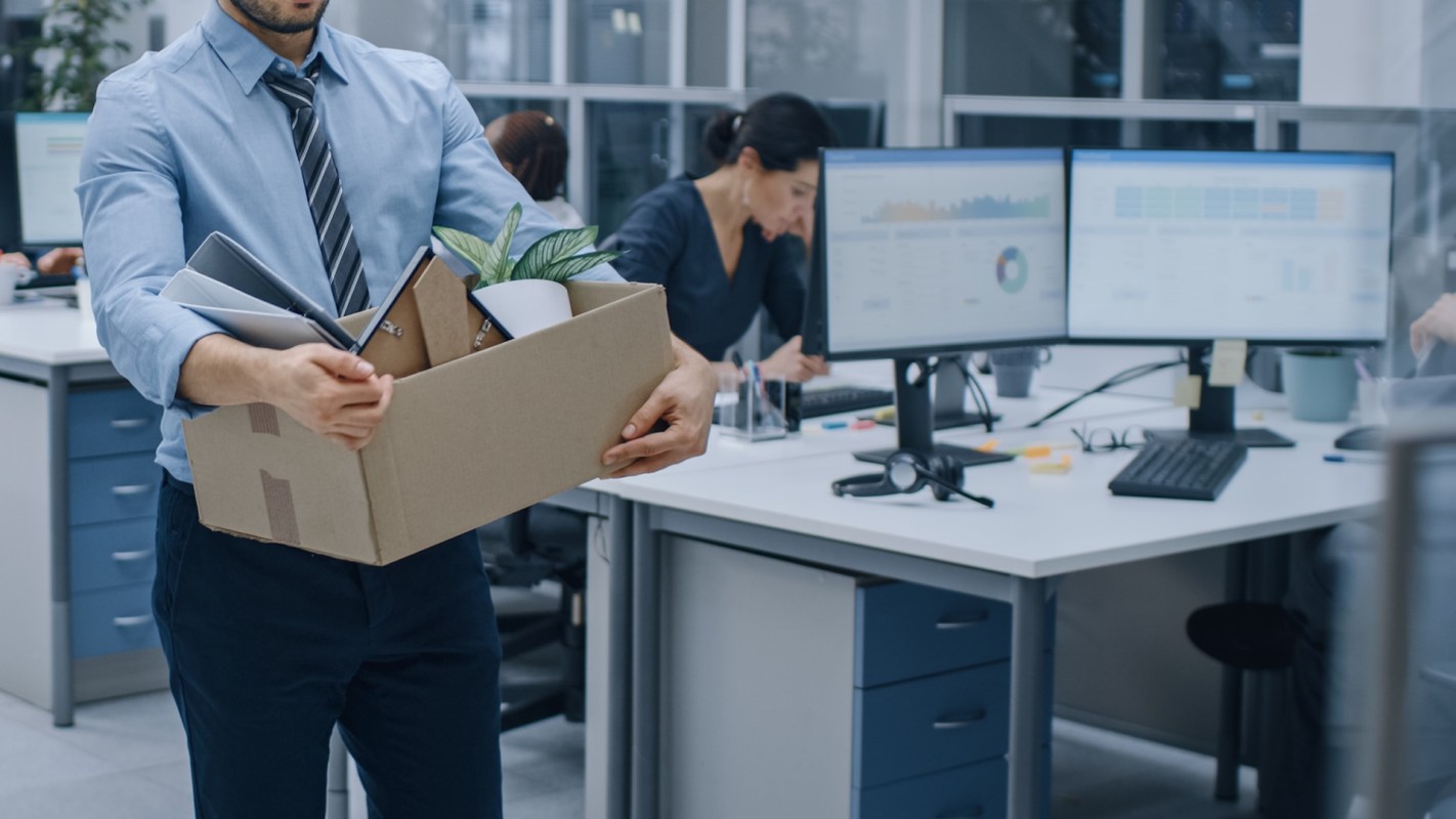 A man holds a box with personal belongings after getting laid off while colleagues work at desks with computer monitors.