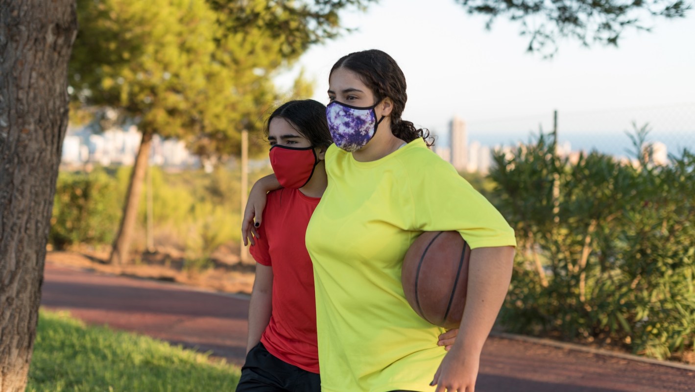 Two girls wearing masks, one in a red shirt and the other in a yellow shirt, stroll together holding a basketball.