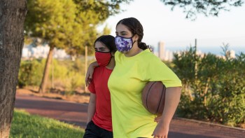 Two girls wearing masks, one in a red shirt and the other in a yellow shirt, stroll together holding a basketball.