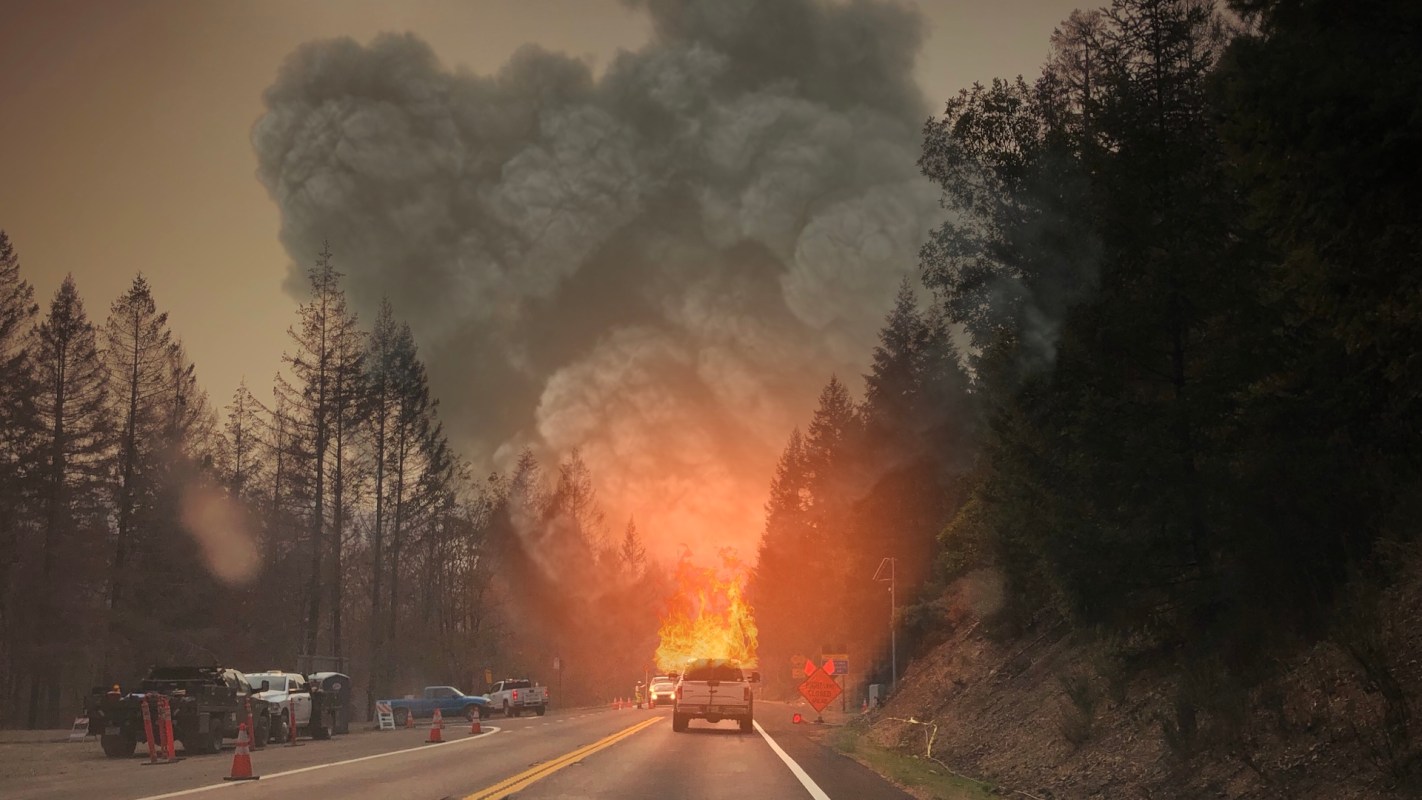 A highway scene with smoke and flames rising from a forest fire, surrounded by emergency vehicles and traffic cones.