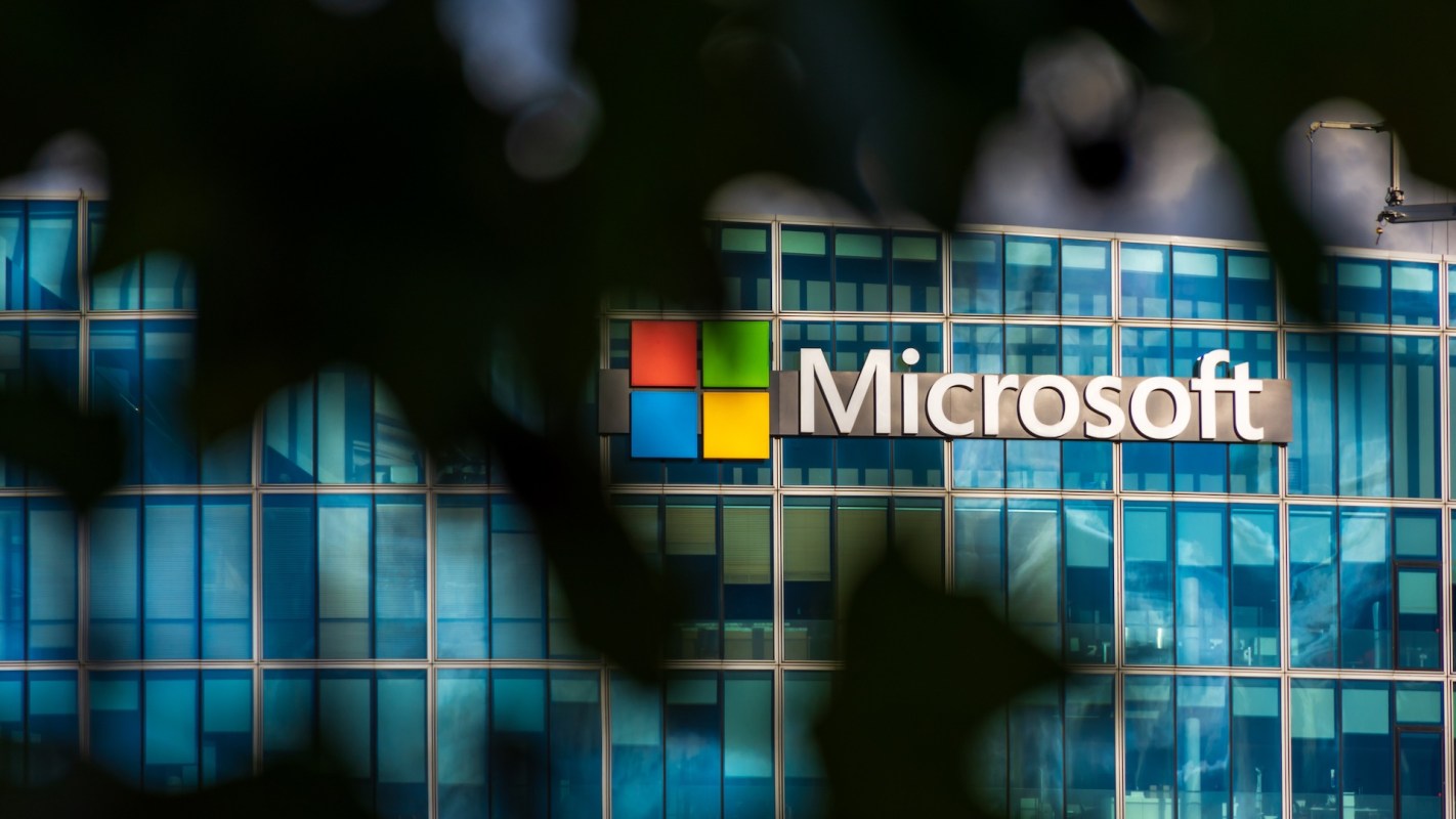 The Microsoft logo is displayed on a modern glass building surrounded by green foliage.