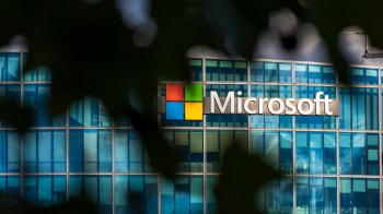 The Microsoft logo is displayed on a modern glass building surrounded by green foliage.