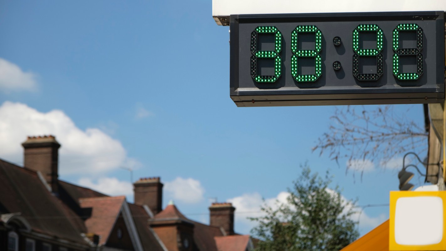 A digital thermostat displaying 38 degrees Celsius against a clear blue sky with clouds and rooftops in the background.