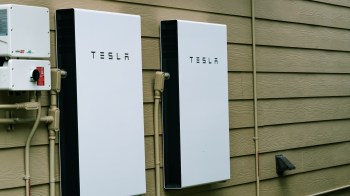 Two Tesla Powerwall battery units mounted on an exterior wooden wall, with an electrical panel nearby.
