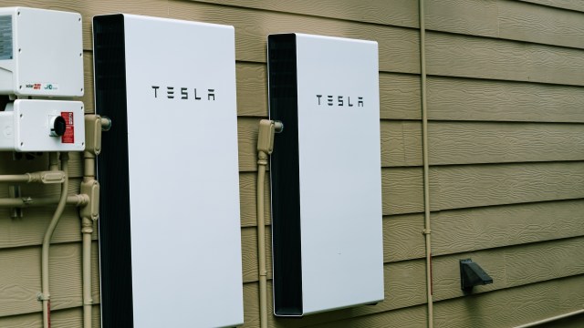 Two Tesla Powerwall battery units mounted on an exterior wooden wall, with an electrical panel nearby.
