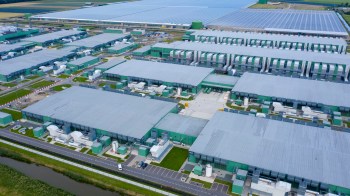 An aerial view of a large industrial complex with green-roofed buildings and solar panels in the background.
