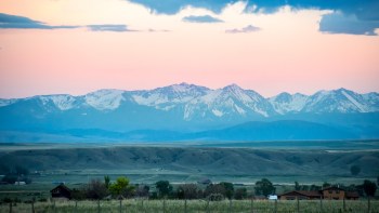 A panoramic view of snow-capped mountains under a pastel sunset sky, with green valleys and scattered houses in the foreground.