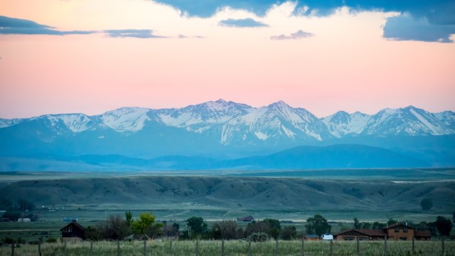 A panoramic view of snow-capped mountains under a pastel sunset sky, with green valleys and scattered houses in the foreground.