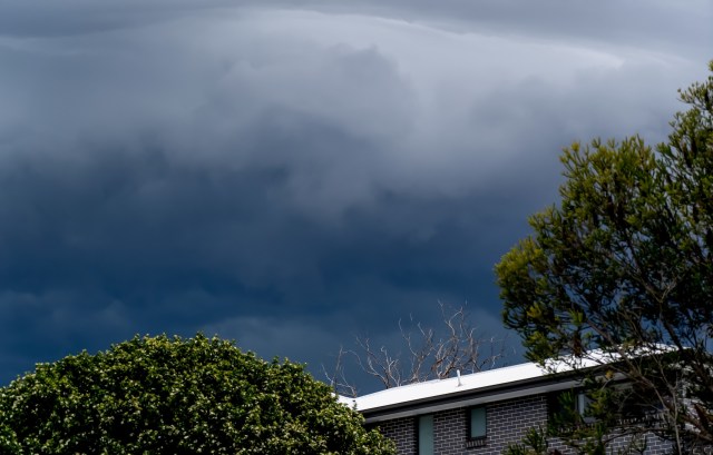 A dark, cloudy sky looms over a residential building and lush green trees.