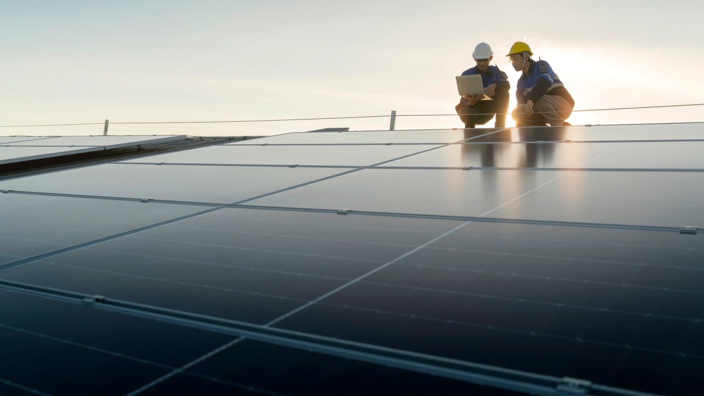 Two technicians in hard hats are inspecting solar panels on a rooftop while using a laptop.