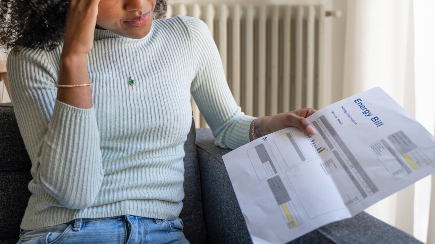 A woman sitting on a couch looks stressed while reading her energy bill.