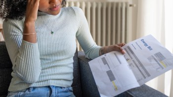 A woman sitting on a couch looks stressed while reading her energy bill.