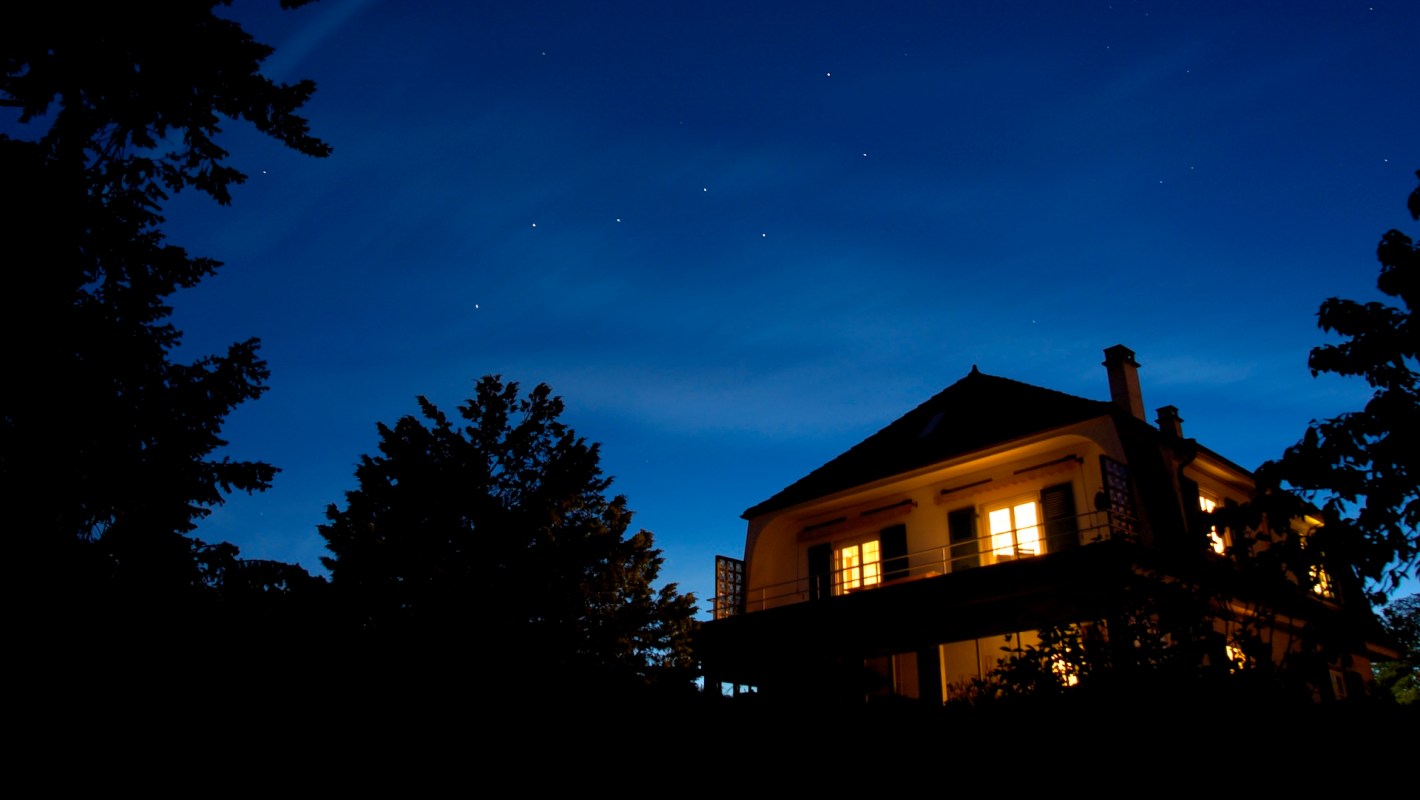 A two-story house with illuminated windows silhouetted against a dark blue sky filled with stars.