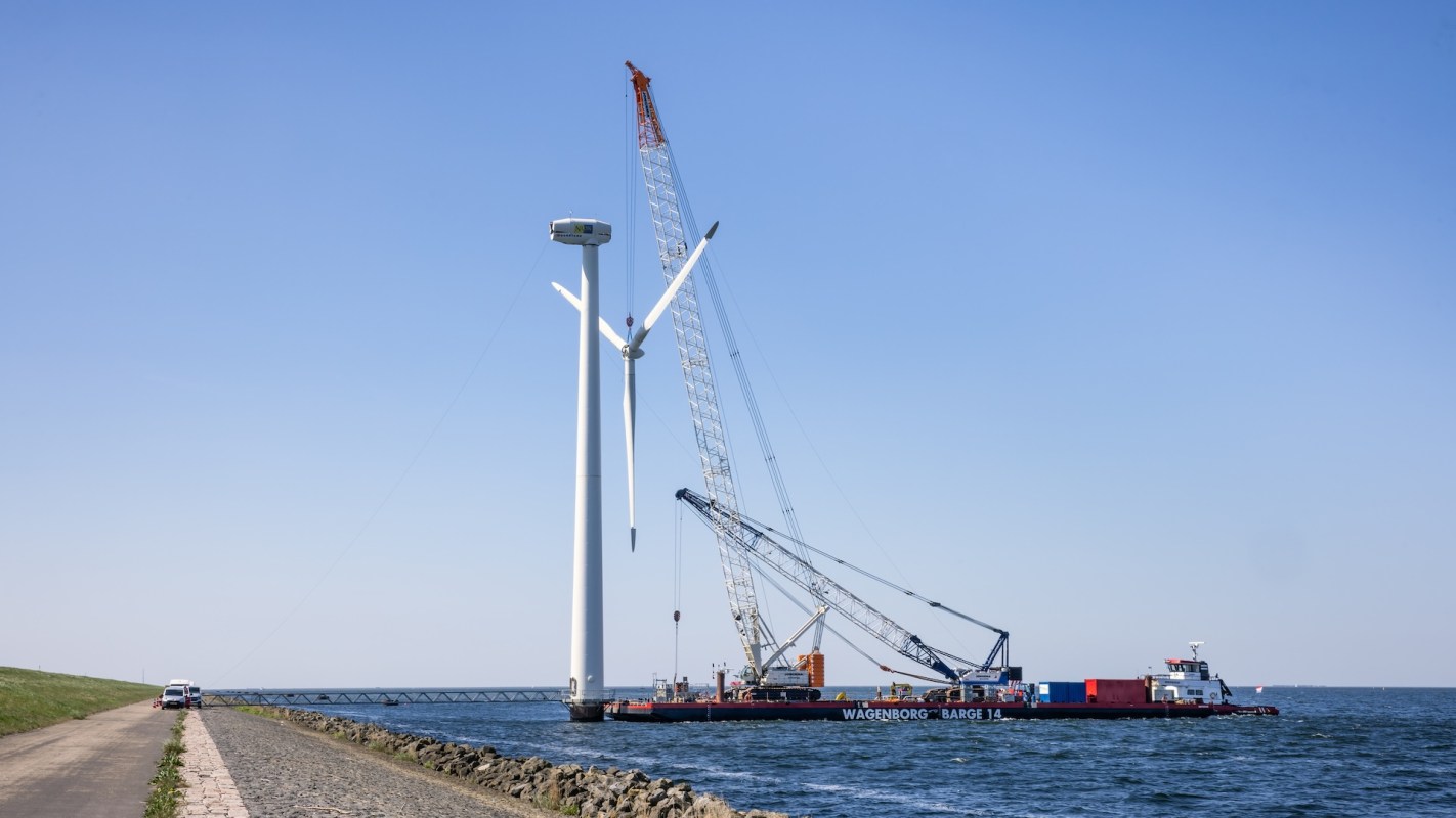A crane-equipped barge is installing a wind turbine over water, next to a road and a grassy hill.