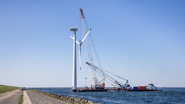 A crane-equipped barge is installing a wind turbine over water, next to a road and a grassy hill.