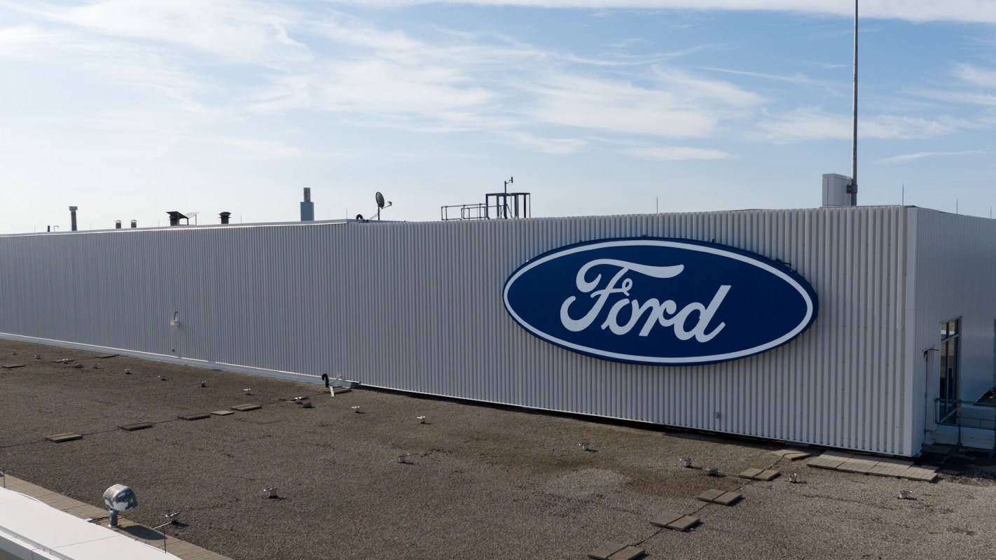 A close-up view of a Ford logo on the side of a white industrial building against a clear blue sky.