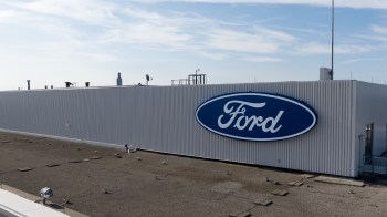 A close-up view of a Ford logo on the side of a white industrial building against a clear blue sky.