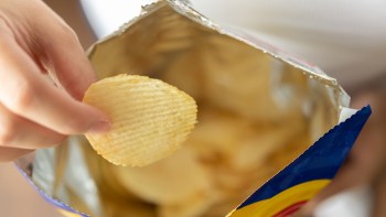 A hand holding a potato chip above an open bag filled with more chips.