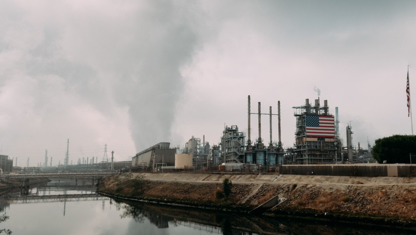 An oil refinery with smoke coming out its stacks and adorned with an American flag, under a cloudy sky.