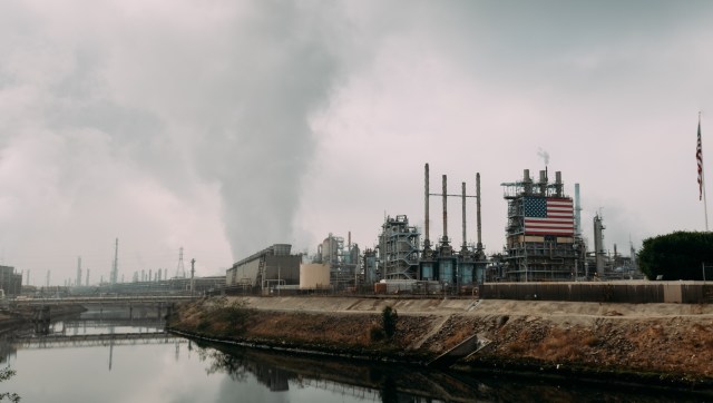 An oil refinery with smoke coming out its stacks and adorned with an American flag, under a cloudy sky.