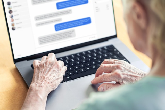 An older adult typing on a laptop while engaged in a messaging conversation on the screen.