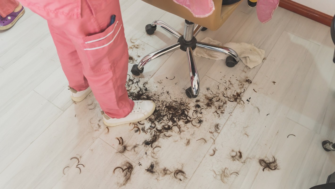 A person in pink pants stands over a floor scattered with dark hair clippings in a salon setting.