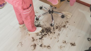 A person in pink pants stands over a floor scattered with dark hair clippings in a salon setting.