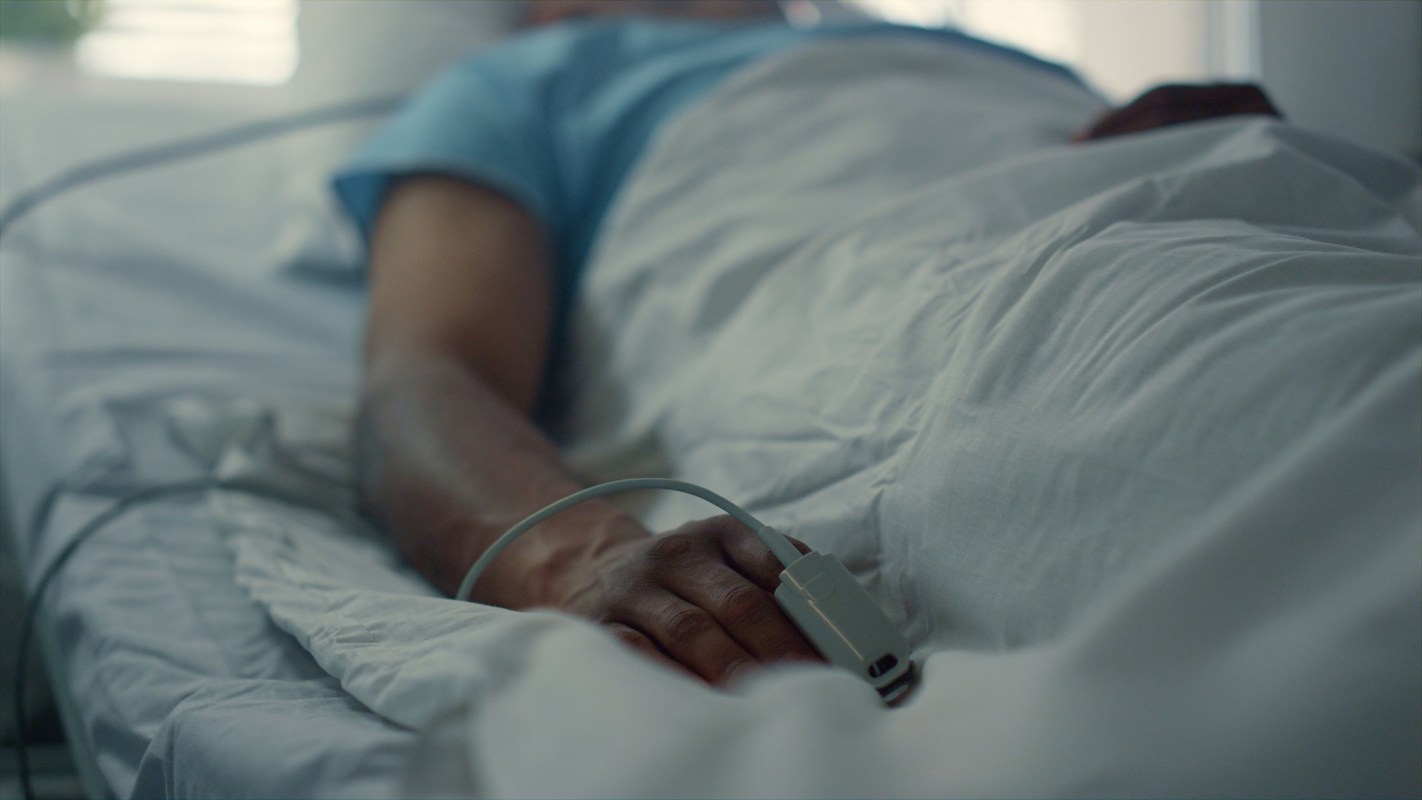 A close-up of a person's hand resting on a hospital bed in a darkened room with medical equipment attached.