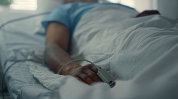 A close-up of a person's hand resting on a hospital bed in a darkened room with medical equipment attached.
