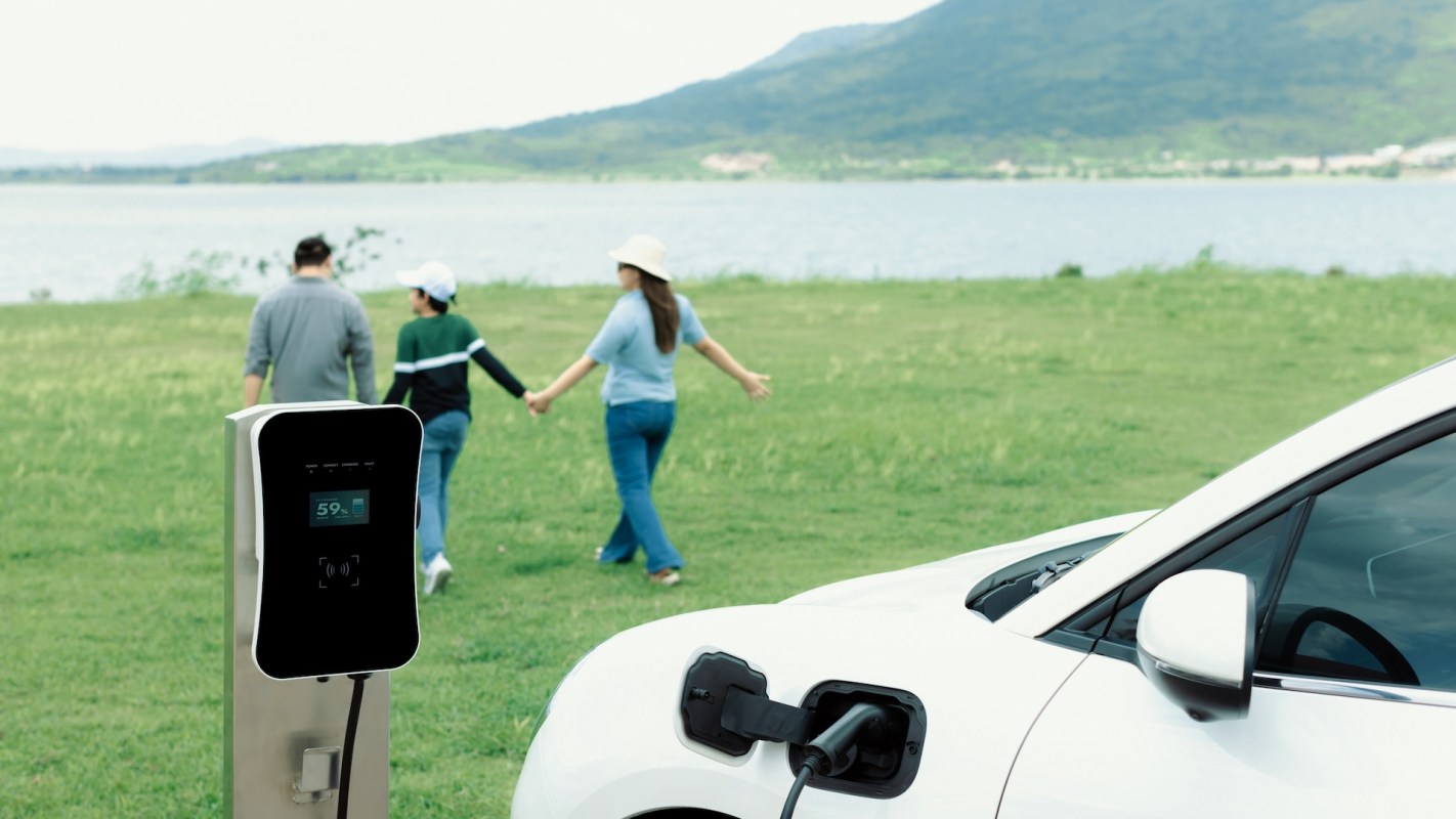 An electric vehicle is charging at a station while three people walk hand-in-hand beside a lake.