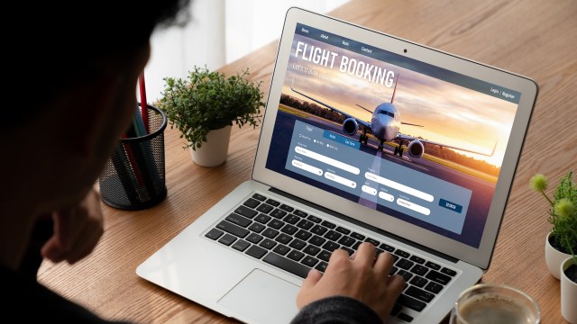 A person booking a flight on a laptop with a travel website displayed, surrounded by plants and stationery.