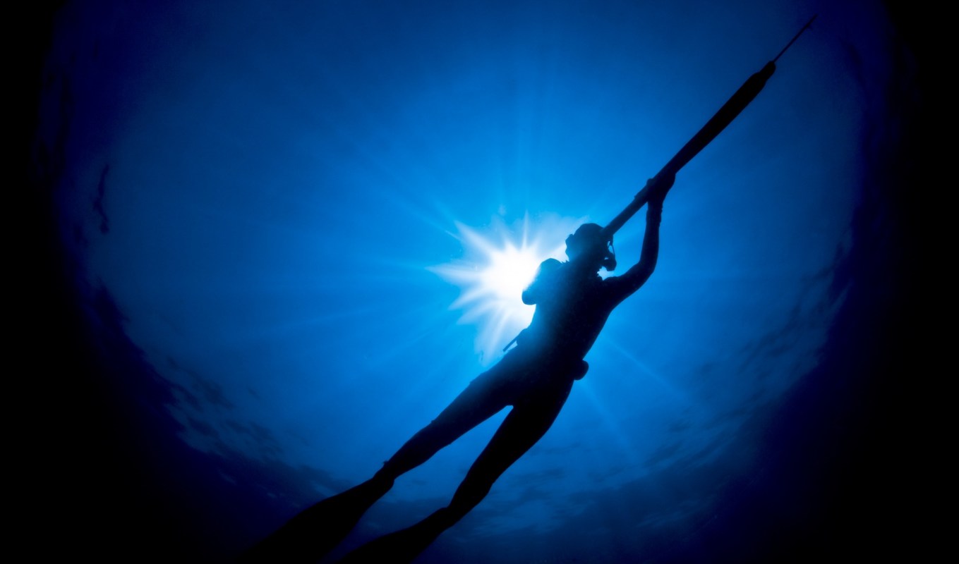 A silhouette of a diver holding a spear gun against a bright blue underwater backdrop with sunlight rays.