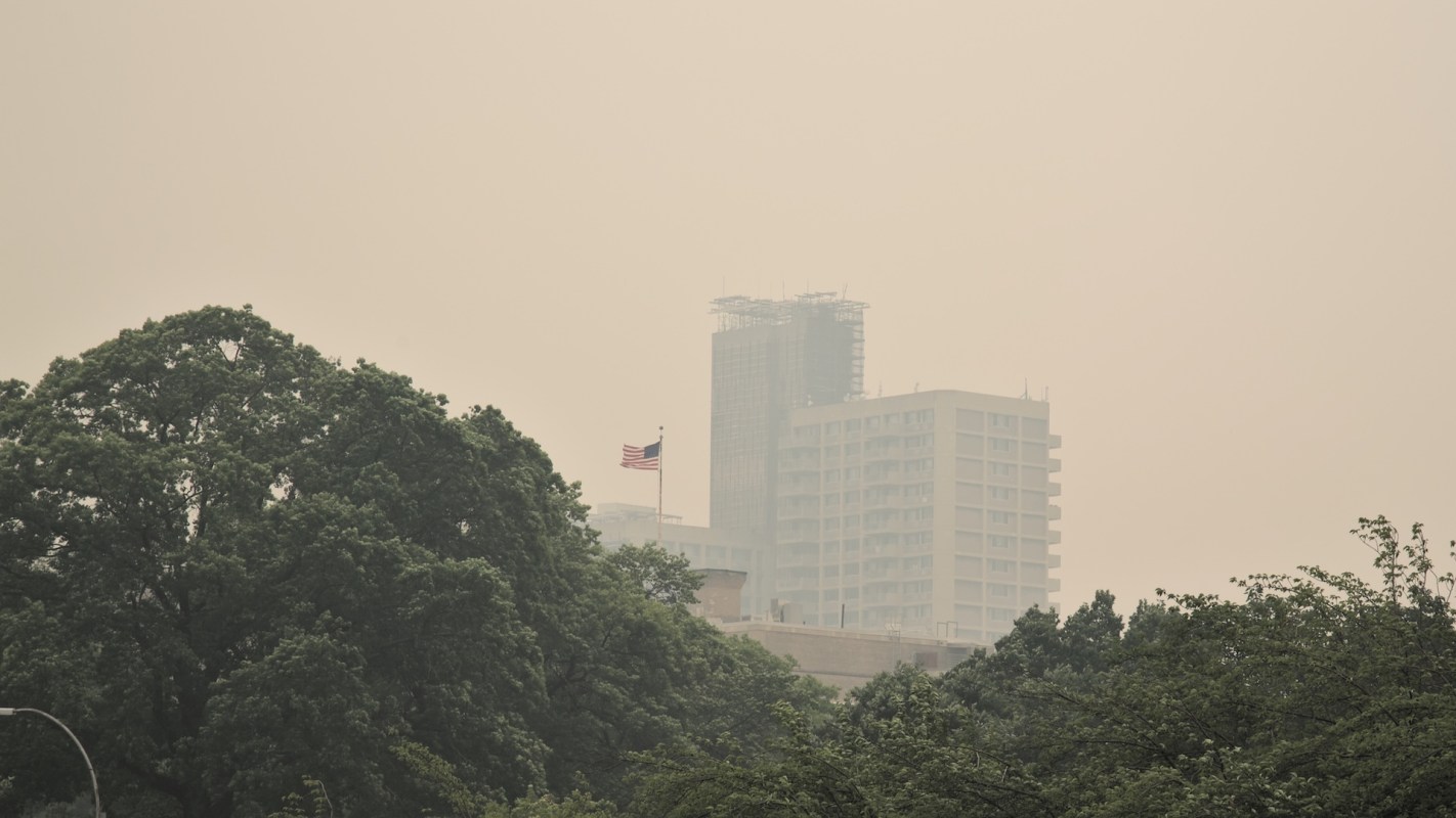A cityscape shrouded in haze, featuring tall buildings and an American flag visible amid green trees.
