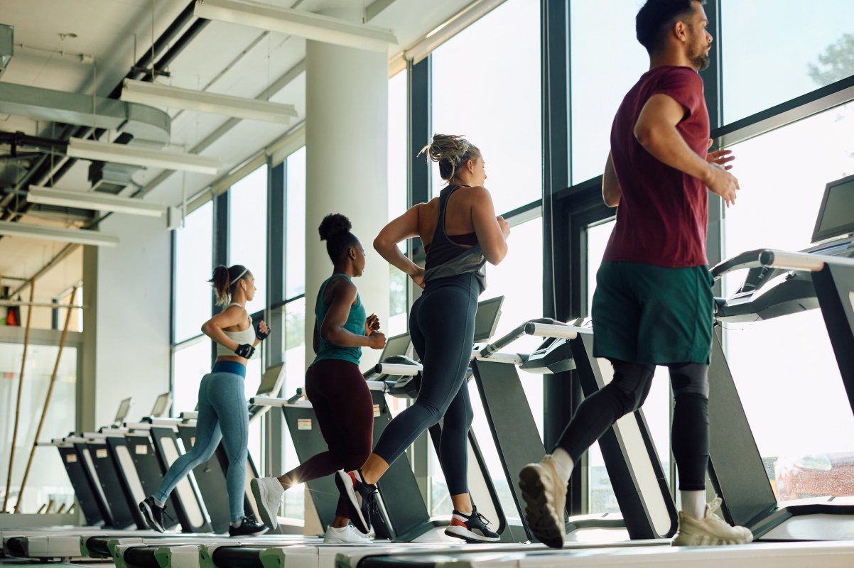 Four individuals are jogging on treadmills in a bright gym with large windows.