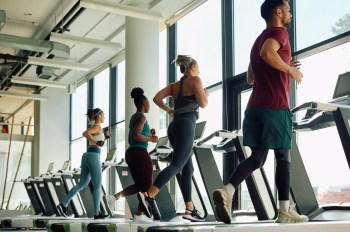 Four individuals are jogging on treadmills in a bright gym with large windows.