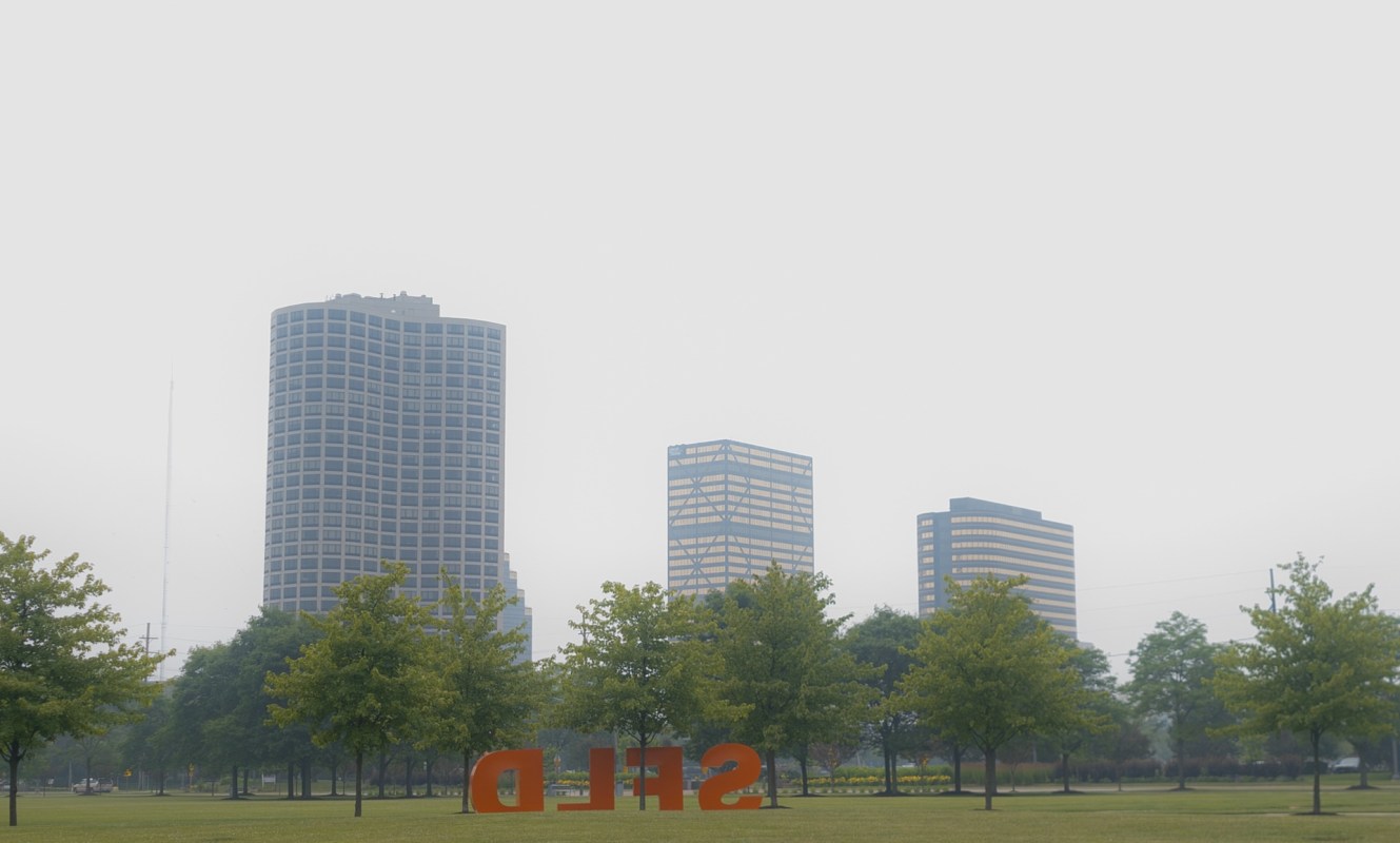 A urban landscape featuring smog over tall buildings and trees, with an orange sculpture in the foreground.