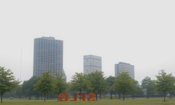 A urban landscape featuring smog over tall buildings and trees, with an orange sculpture in the foreground.