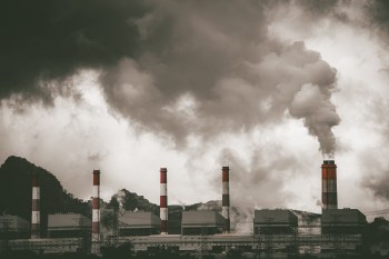 A coal power plant with smokestacks releasing thick smoke against a backdrop of dark, cloudy skies.
