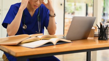 A stressed medical student in scrubs looks at a laptop with open books on a wooden desk.