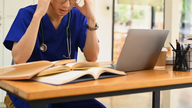 A stressed medical student in scrubs looks at a laptop with open books on a wooden desk.