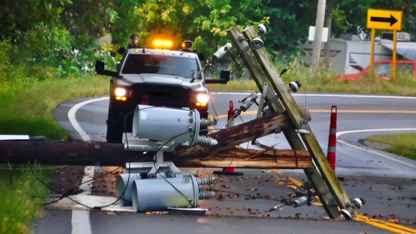 A downed utility pole with equipment on the road alongside a vehicle and warning cones.