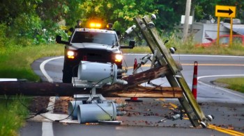 A downed utility pole with equipment on the road alongside a vehicle and warning cones.