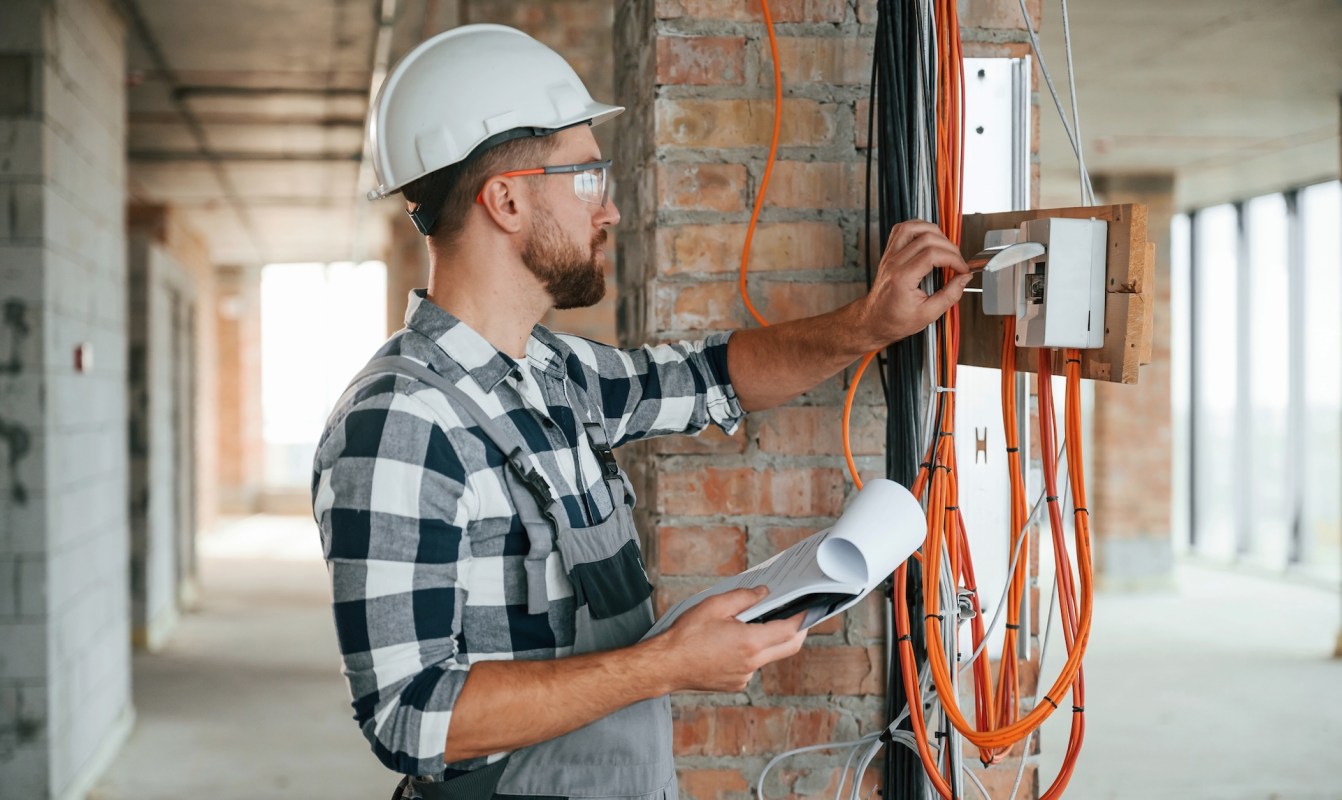 A construction worker in a hard hat examines wiring while holding a clipboard.