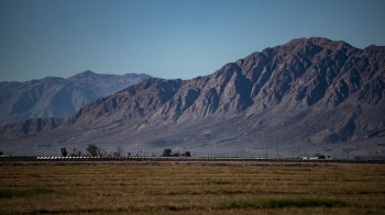 Vast landscape featuring rugged mountains in the background and flat farmland in the foreground under a clear blue sky.