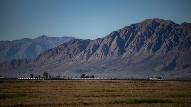 Vast landscape featuring rugged mountains in the background and flat farmland in the foreground under a clear blue sky.